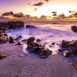 Surf washing between jagged shoreline rocks beneath a warm, cloud-filled sky