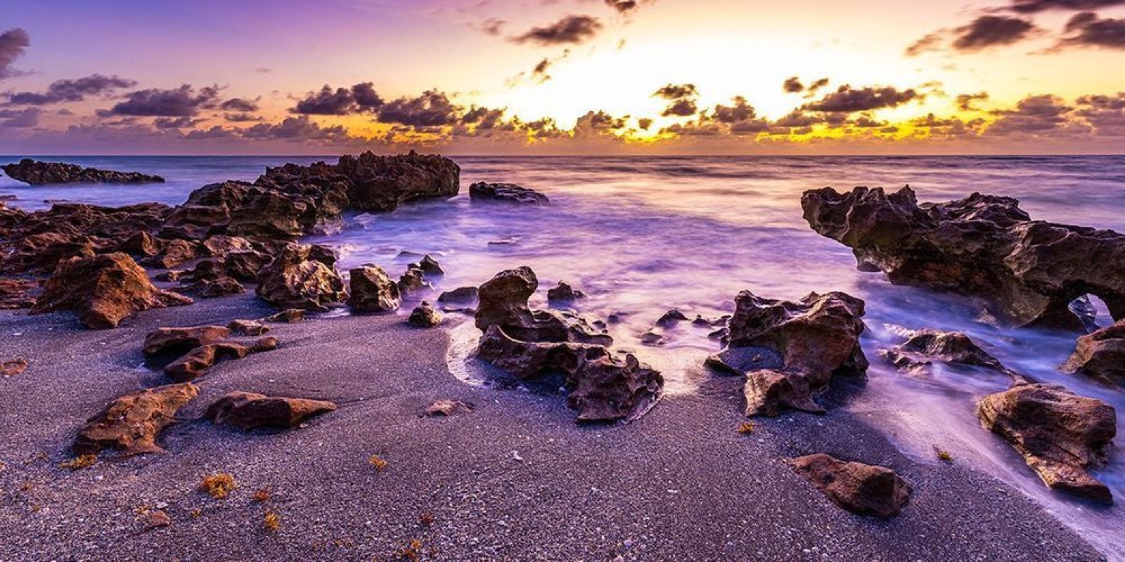 Surf washing between jagged shoreline rocks beneath a warm, cloud-filled sky