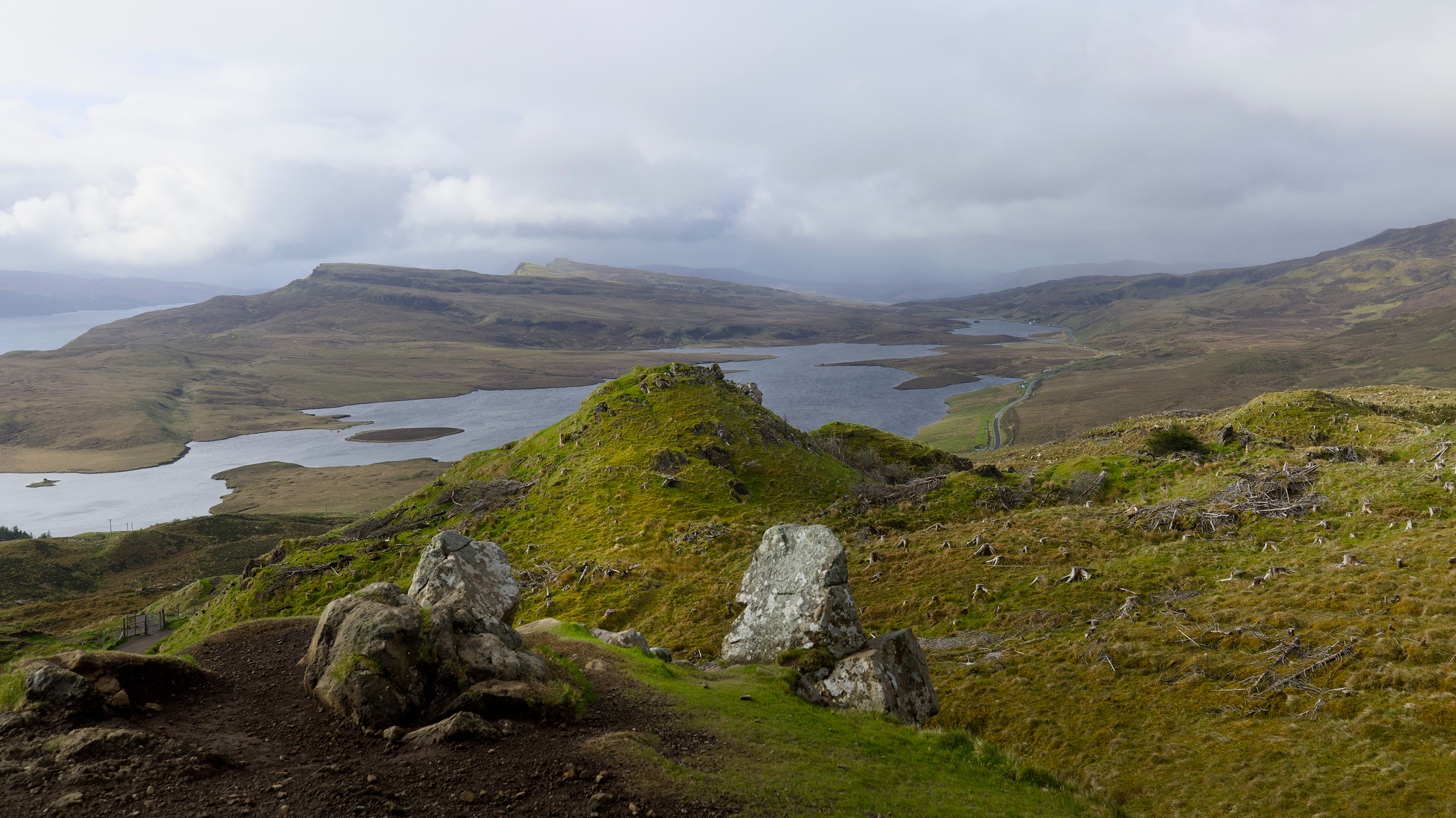 Rocky green hillside at The Storr overlooking lochs and distant ridges beneath a cloudy sky.